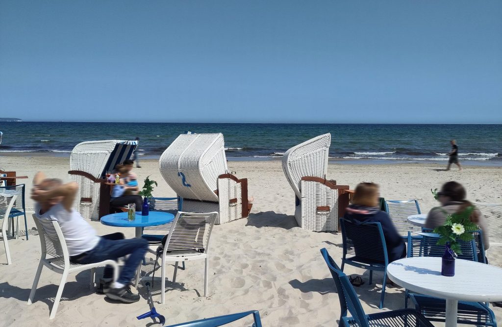Bild von einem Strandcafé mit blick auf das Meer. Im Vordergrund sind weiße und blaue runde Tische mit Gartenstühlen zu sehen. Dahinter stehen drei Strandkörbe. Auf zwei Tischen sind Blumen aus Plastik zu sehen. Der Himmel ist wolkenlos und blau. 